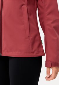Giacca softshell bordeaux con zip anteriore, tasche laterali e chiusure ai polsi con velcro, abbinata a pantaloni neri aderenti. Texture liscia.