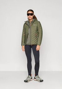 Lekka oliwkowozielona kurtka puchowa na zamek, z elastycznymi mankietami i pomarańczowymi akcentami, noszona na szarym topie i czarnych legginsach, do kompletu szare buty.