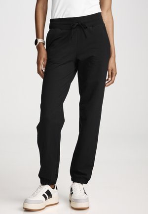 Person trägt schwarze Jogginghose mit Kordelzug und elastischen Bündchen sowie weiße Sneakers mit schwarzen Streifen und steht vor weißem Hintergrund.