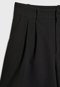 Pantalon noir à fines rayures, confectionné sur mesure, avec plis, passants de ceinture et poches latérales, montré de la taille jusqu’à mi-cuisse.