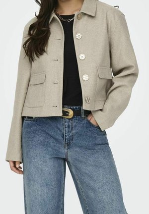 Femme portant une veste beige boutonnée par-dessus un haut noir, avec un jean bleu taille haute et une ceinture noire avec une boucle dorée.