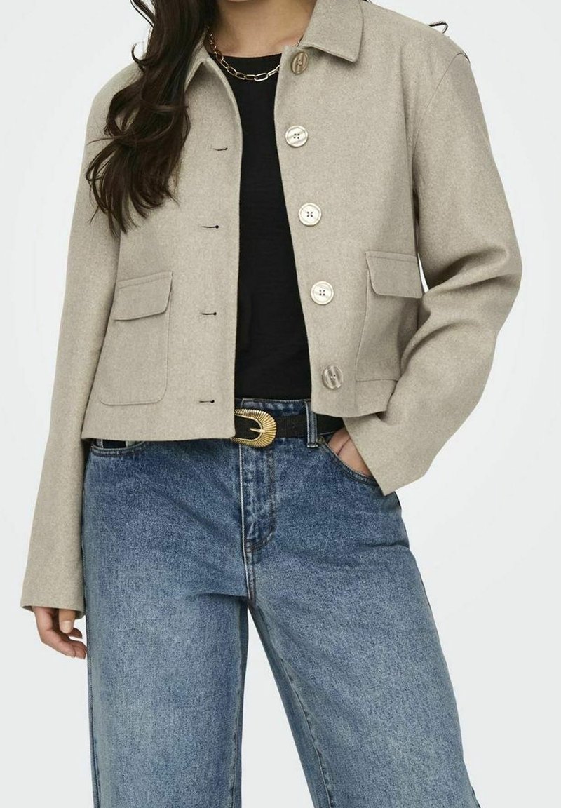 Femme portant une veste beige boutonnée par-dessus un haut noir, avec un jean bleu taille haute et une ceinture noire avec une boucle dorée.