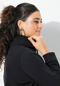 Sudadera negra con capucha y alto cuello, con una textura suave. Modelo llevando grandes pendientes de aro retorcidos y con el pelo ondulado recogido en una coleta.