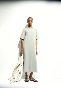 Hellgraues, knielanges Kleid mit kurzen Ärmeln; weiche Textur. Kombiniert mit Sandalen mit schwarzem Grund und grünen Akzenten. Hält einen beigen Schal.