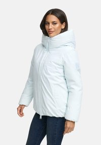 Donna che indossa un giubbotto imbottito azzurro con colletto alto e zip frontale, abbinato a jeans blu scuro, in piedi davanti a uno sfondo neutro.
