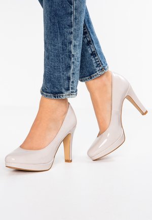 Escarpins à talons hauts en cuir verni beige clair usé, associés à un jean en denim bleu, montrant les pieds et le bas des jambes sur un fond blanc.