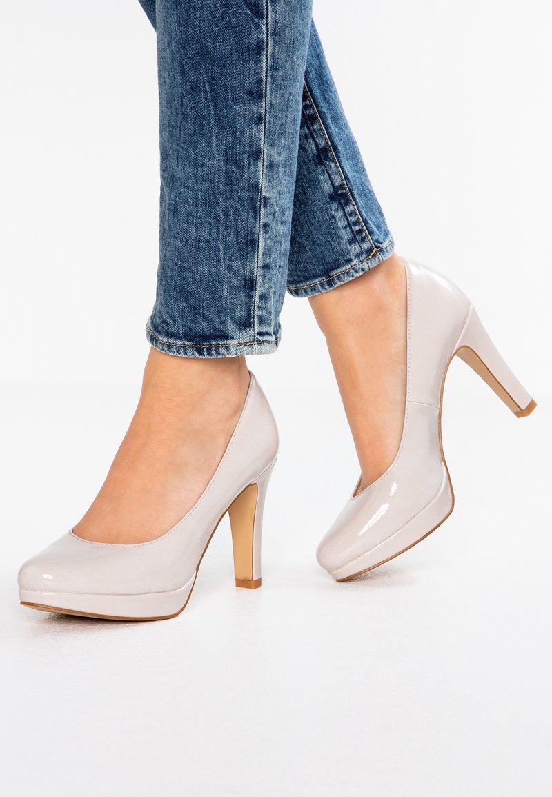 Escarpins à talons hauts en cuir verni beige clair usé, associés à un jean en denim bleu, montrant les pieds et le bas des jambes sur un fond blanc.