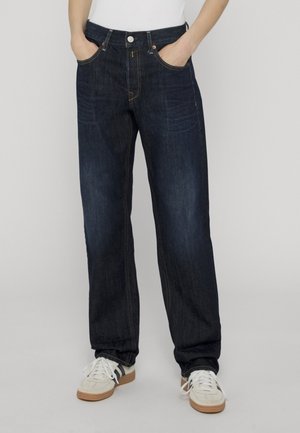 Person iført mørkeblå afslappede jeans med hænderne i lommerne og hvide sneakers med gummisåler på en ensfarvet baggrund.
