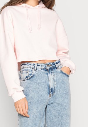 Vrouw draagt een korte, lichtroze hoodie en een lichtblauwe, hooggetailleerde jeans met acid-was-effect, met één hand in de zak.