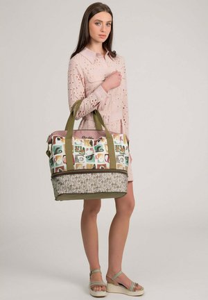 Junge Frau in einem hellrosa Kleid mit Lochmuster, die eine große, mehrfarbig gemusterte Tragetasche mit olivgrünen Trägern hält.
