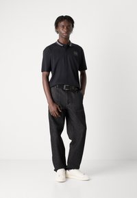 Zwarte polo met grijze kraagdetails, gecombineerd met losse donkere denim jeans en witte sneakers. Kenmerken zijn zichtbare stiknaden en een riem.