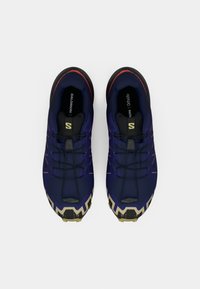 Sapatos de desporto roxos e pretos com uma sola de borracha, apresentando uma parte superior texturizada, um sistema de atacadores proeminente e um padrão em zig-zag na sola.