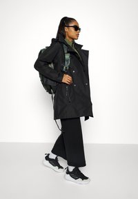 Czarny wodoodporny płaszcz trench z podwójnym zapięciem, noszony z zielonym plecakiem w kamuflażu, czarnymi spodniami typu wide-leg i czarnymi butami sportowymi.