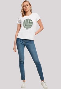 Weißes T-Shirt mit einem grünen gestreiften, kreisförmigen grafischen Motiv. Kombiniert mit blauen Skinny-Jeans und weißen Turnschuhen. Das Model steht vor einem neutralen Hintergrund.