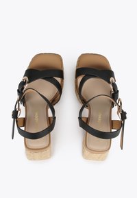 Sandalias de plataforma negras con suela de yute trenzado, que presentan correas entrelazadas y un detalle de hebilla en oro rosa. Plantilla de color marrón suave.