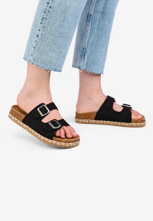 Black suede sandals with two adjustable straps, silver hardware, and a braided jute sole, paired with cropped light blue jeans.