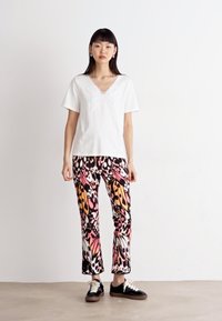 Weißes Kurzarmoberteil mit dekorativem Ausschnitt, kombiniert mit gemusterten Hosen mit pinken, orangen und schwarzen Blumenmustern. Schwarze Sneakers.
