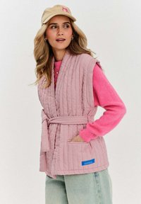 Różowa pikowana kamizelka z paskiem w talii i kieszeniami, noszona na różowym swetrze. Posiada fakturowany materiał i niebieską metkę marki. Casualowy, lekki design.