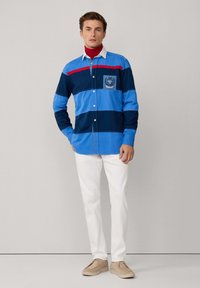 Lange mouw overhemd met blauwe en marine gestreepte patroon en een rode accenten, met een borstzak en geborduurd logo, gecombineerd met witte broek.