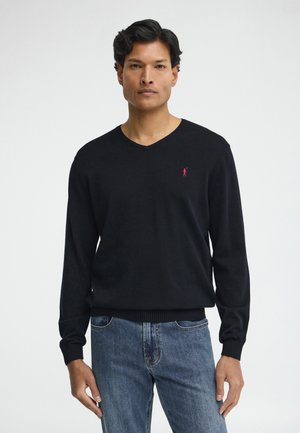 Mann trägt schwarzen V-Ausschnitt-Pullover mit kleinem rotem Logo auf der Brust und blaue Jeans vor schlichtem weißem Hintergrund.