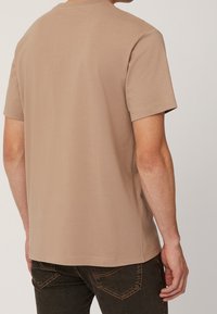 Beige bomulls t-shirt med kort ärm och rund halsringning. Slät textur; nederkanten vilar vid midjan. Minimalistisk design, inga synliga mönster.