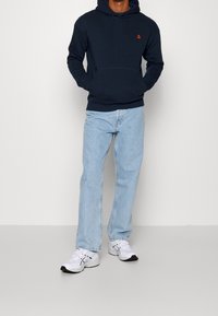 Sudadera con capucha azul marino, con un bolsillo delantero y un logo rojo, combinada con jeans rectos de mezclilla azul claro y zapatillas deportivas blancas con detalles en negro.