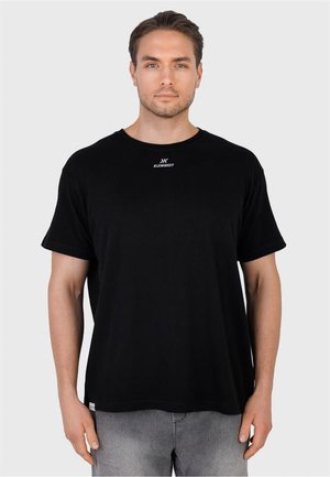 Hombre con camiseta negra lisa con un pequeño logo blanco en el centro del pecho, combinado con pantalones grises, de pie frente a un fondo liso.