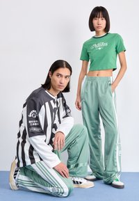 Camisa de mangas longas cinza e branca às riscas; t-shirt curta verde; calças de treino verdes claras com riscas brancas; ambos usam ténis com detalhes.