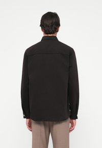 Crna košulja dugih rukava s ovratnikom, dugmadima na mansetama i ravnim rubom. Izrađena od čvrstog materijala, s čistim, minimalističkim dizajnom.