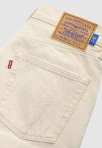 Κοντινή λήψη πίσω τσέπης μπεζ τζιν Levi's με κόκκινη ετικέτα Levi's και δερμάτινη ετικέτα Levi Strauss & Co. σε ταμπά χρώμα που δείχνει το μέγεθος W32 L.