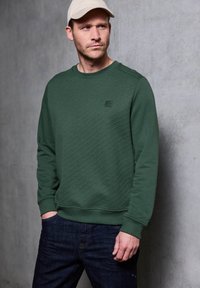 Felpa verde con un motivo diagonale testurizzato, collo rotondo e polsini a costine, abbinata a jeans di denim blu scuro e un cappellino di colore chiaro.