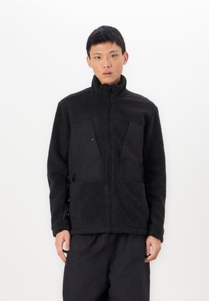 Chaqueta de forro polar negra con un cuello alto, que presenta paneles de tela negra contrastantes en el pecho y bolsillos con cremallera. Material suave y texturizado.