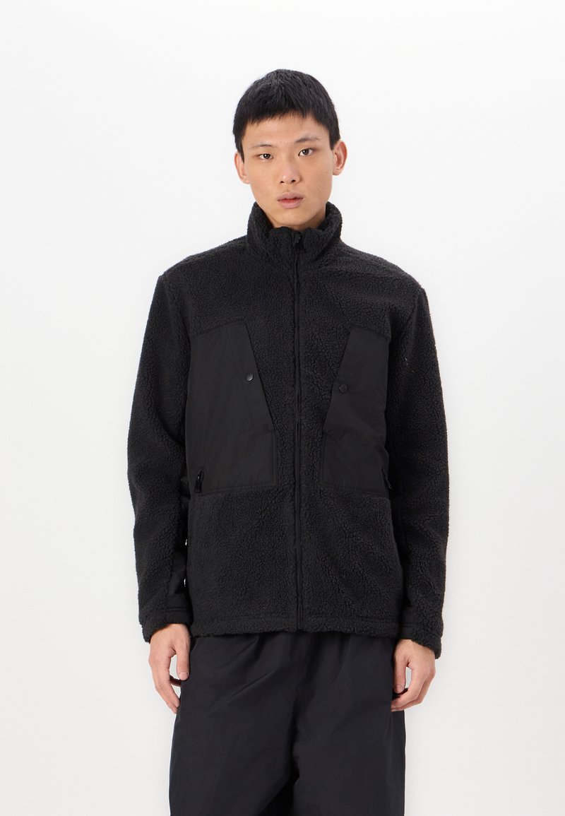 Schwarze Fleecejacke mit hohem Kragen, mit kontrastierenden schwarzen Stoffpaneelen auf der Brust und mit Reißverschlüssen versehenen Taschen. Weiches, strukturiertes Material.