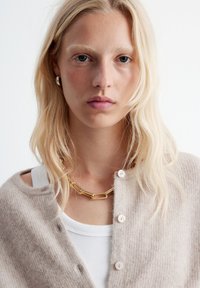 Beige Strickjacke mit Knöpfen, über einem weißen Tanktop getragen. Akzentuiert mit einer goldenen Halskette. Weiche Textur und einfaches Design.