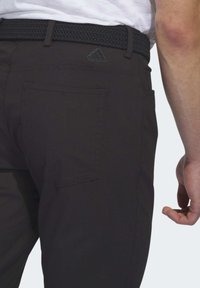 Sorte bukser med baglomme og vævet bælte båret med hvid skjorte, som viser en del af højre arm og hånd mod en lys baggrund.