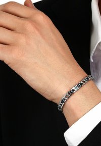 Metal bracelet with alternating black and silver links, featuring a central logo detail, worn on a hand with a textured, natural finish.