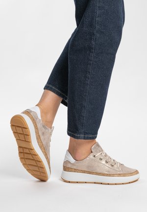 Lässige beige Sneaker mit Kork-Details und weißen Sohlen, getragen zu dunkelblauen, gekürzten Jeans, auf einem erhöhten Fuß vor weißem Hintergrund.