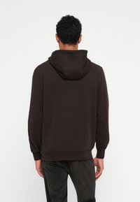 Καφέ φούτερ κατασκευασμένο από μαλακό ύφασμα, με κουκούλα με κορδόνι, πλεγμένους καρπούς και χαλαρή εφαρμογή. Η πίσω όψη αποκαλύπτει ελ�άχιστες λεπτομέρειες.