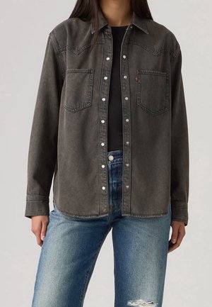 Personne portant une chemise en jean noire délavée à boutons sur un top noir avec un jean bleu usé.