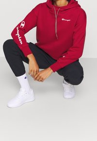 Czerwona bluza z kapturem z białym logo "Champion" z przodu i na rękawie, w parze z czarnymi dresami i białymi trampkami.