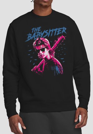 Mann trägt schwarzen Pullover mit pink-blauem Motiv einer Person, die eine Stachelkeule hält, und dem Text "The Babysitter" darüber.