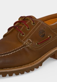 Chaussure bateau en cuir marron avec lacets rouges, coutures blanches, semelle épaisse crantée et logo arbre embossé sur le côté.