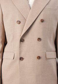 Beige double-breasted blazer with a subtle texture, featuring brown buttons and two side pockets. Tailored design without visible lining.