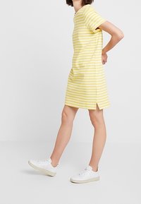 Henkilö, joka pukeutuu kelta-valkoiseen raidalliseen lyhythihaiseen mekkoon ja valkoisiin tennareihin, seisoo toinen jalka eteen ojennettuna.