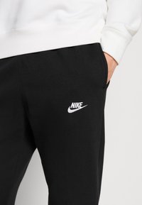 Osoba v černých Nike teplácích s bílým logem a bílém svetru, s rukou v kapse, na �čistém světlém pozadí.