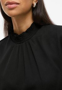 Top nero con un alto collo arricciato e una texture plissè. Accompagnato da un orecchino argento a forma di goccia.