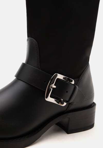 Botte en cuir noir avec un talon de hauteur moyenne, équipée d'une large bride et d'une boucle rectangulaire argentée brillante sur le côté extérieur.