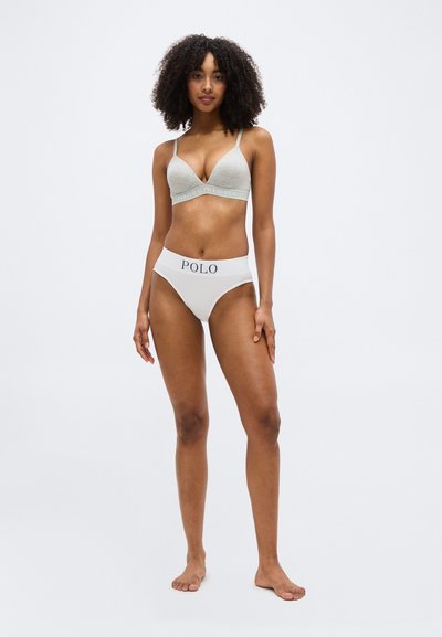 Femme aux cheveux bouclés portant un bralette gris et un sous-vêtement taille haute blanc avec la marque "POLO", debout pieds nus sur un fond blanc.