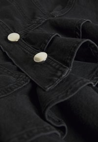 Black denim fabric features visible stitching, silver metal snap buttons, and a ruffled hem detail, creating texture and structure.