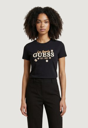 Donna con capelli ricci che indossa una maglietta nera con la scritta "Los Angeles GUESS Est. 1981" e piccoli simboli, abbinata a pantaloni neri.